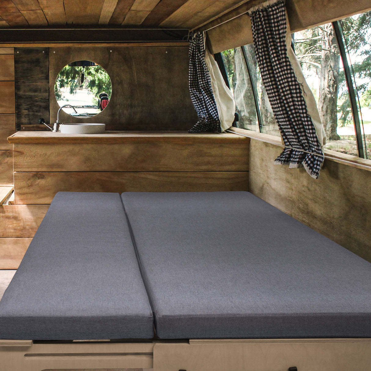 Gemütliches, graues Bett im Camper Van, umgeben von Holzverkleidung und karierten Vorhängen, ideal für entspannte Reisen und Übernachtungen