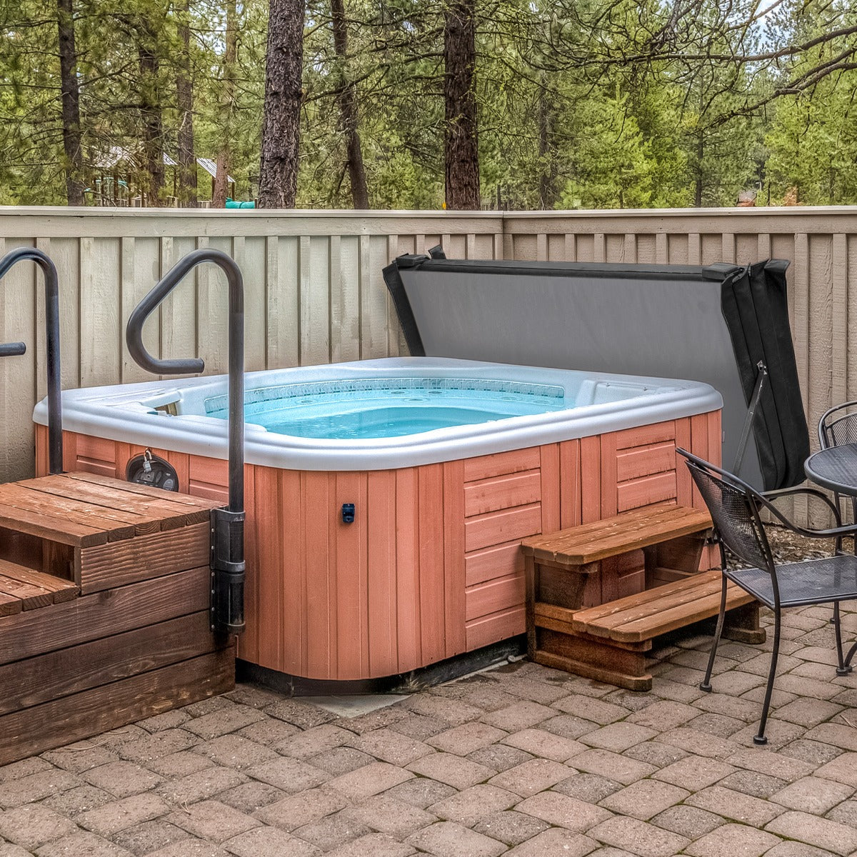Schwarze Whirlpool-Abdeckung auf einem Holz-Whirlpool mit blauen Wasser, umgeben von einem gepflasterten Bereich und Bäumen im Hintergrund