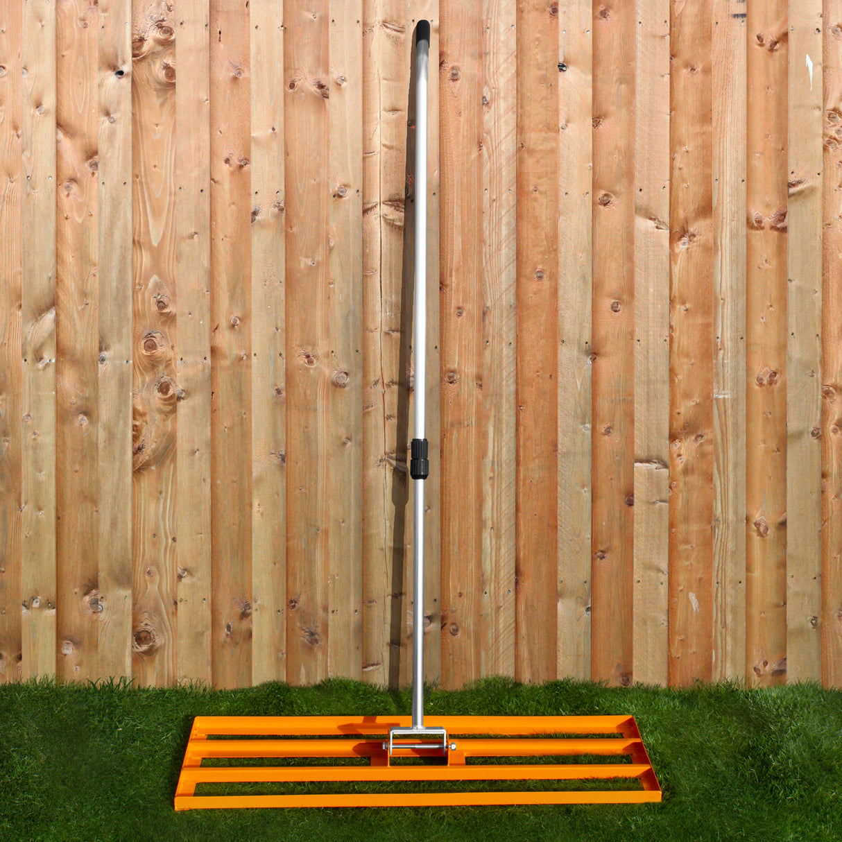 Rasenrakel in leuchtendem Orange mit einem stabilen, silbernen Griff, platziert auf grünem Gras vor einer Holzlattenwand