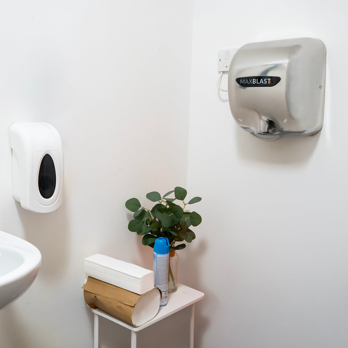 Eleganter MAXBLAST Händetrockner in Silber an der Wand, neben Seifenspender und einer kleinen Pflanze auf einem weißen Tisch