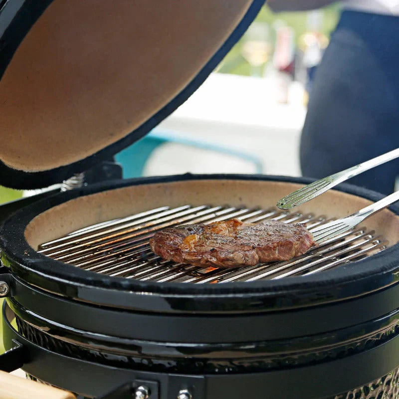 Schwarzer Kamado Holzkohlegrill mit geöffnetem Deckel, auf dem ein saftiges Steak auf den Grillrosten brutzelt, umgeben von einem eleganten Ambiente