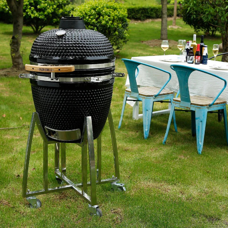 Schwarzer Kamado Holzkohlegrill mit strukturierter Oberfläche, auf einem stabilen Gestell, umgeben von einem eleganten Garten und einem gedeckten Tisch