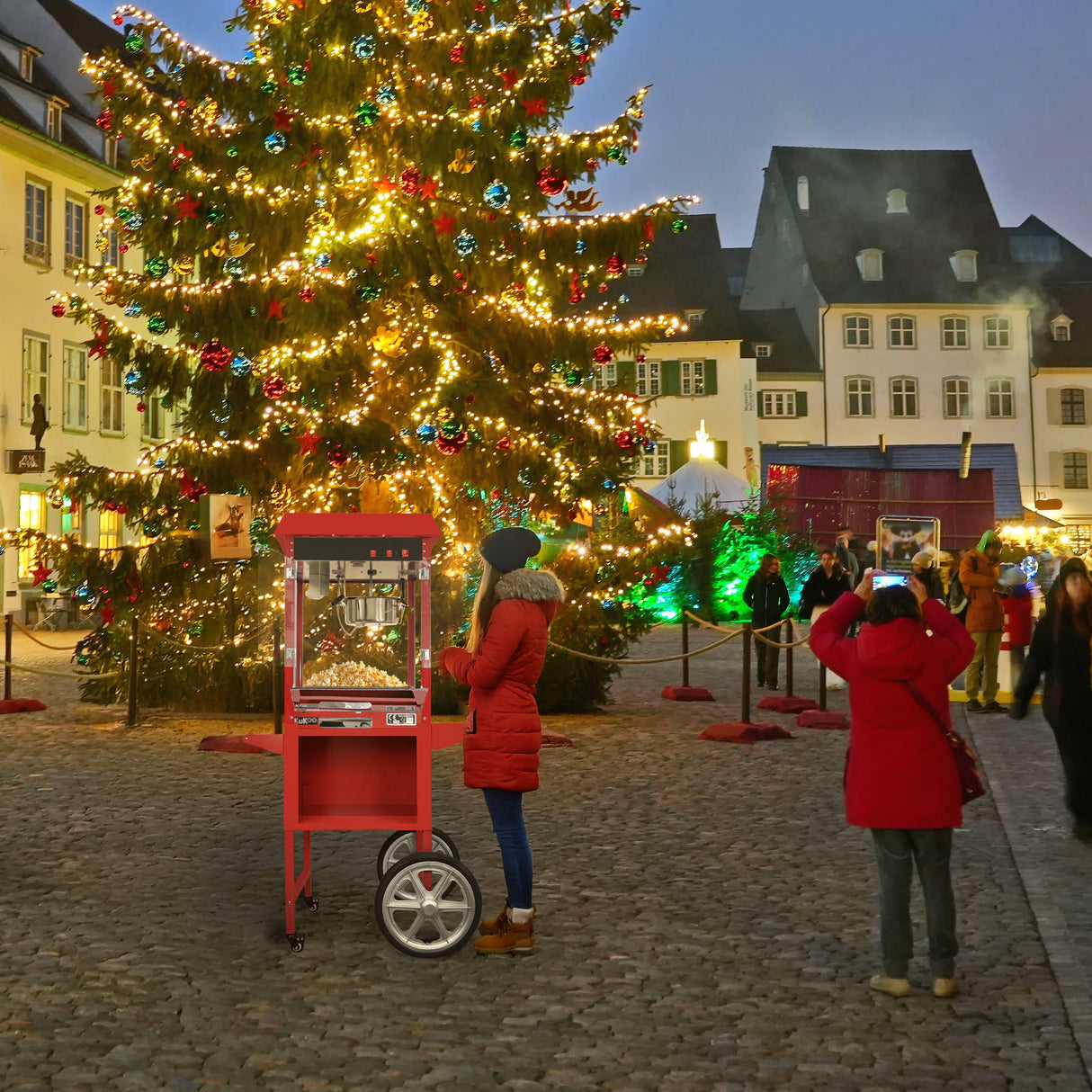 Ein roter KuKoo Popcornmaschinenwagen steht festlich dekoriert vor einem Weihnachtsbaum, umgeben von Menschen in winterlicher Kleidung und bunten Lichtern
