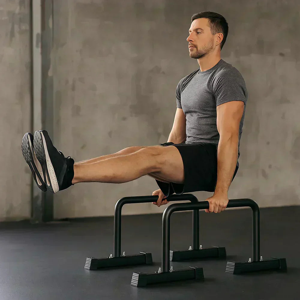 Mann in grauem T-Shirt und Shorts führt einen Beinheber an schwarzen Parallettes in einem minimalistischen Heim-Fitnessstudio aus