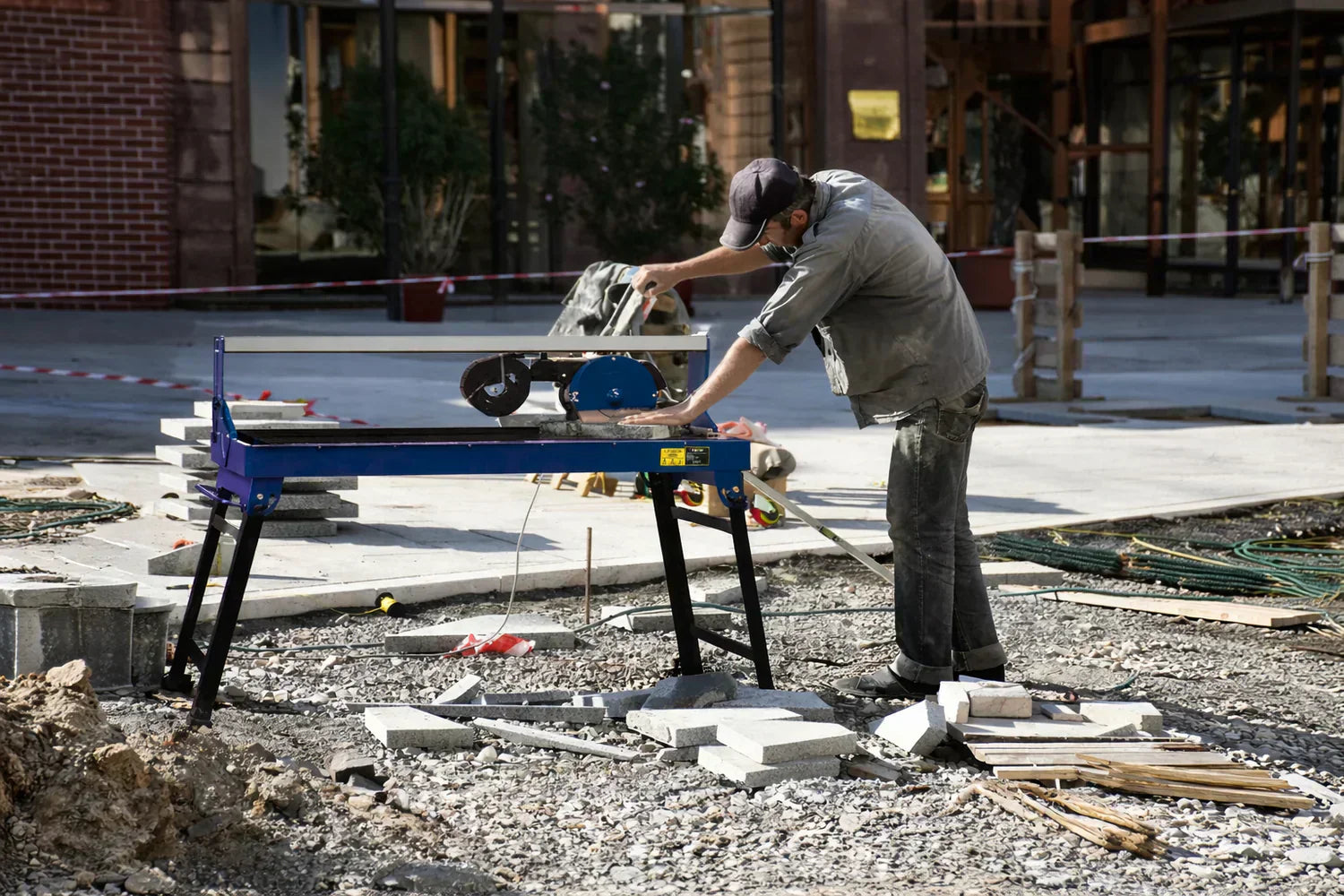 Ein Handwerker schneidet Fliesen mit einem blauen Fliesenschneider auf einer Baustelle, umgeben von Steinen und Holzmaterialien