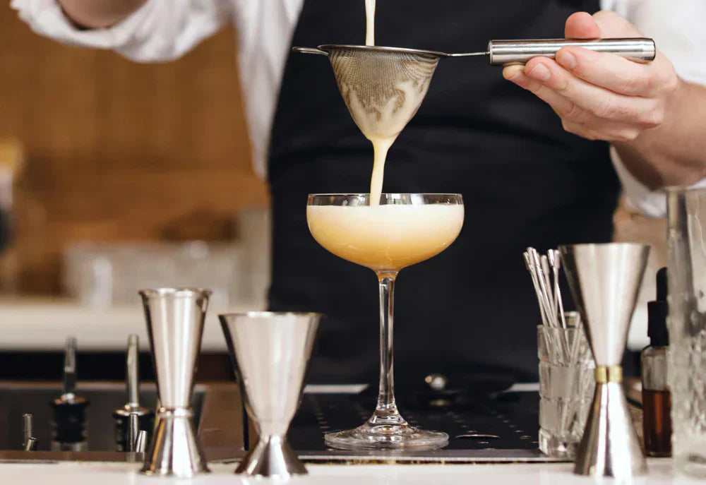 Ein Barkeeper gießt einen cremigen Cocktail durch ein feines Sieb in ein elegantes, hohes Glas, umgeben von Messbechern und Barwerkzeugen