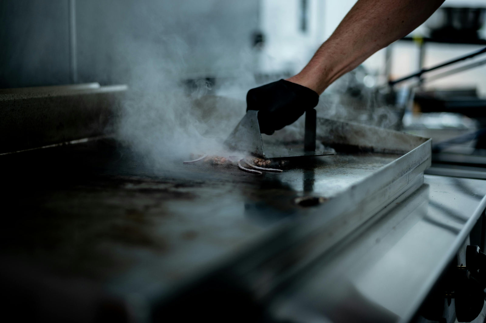 Ein Koch mit schwarzer Handschuhen wendet gegrilltes Fleisch auf einer glänzenden, elektrischen Grillplatte in einer dampfenden Küche
