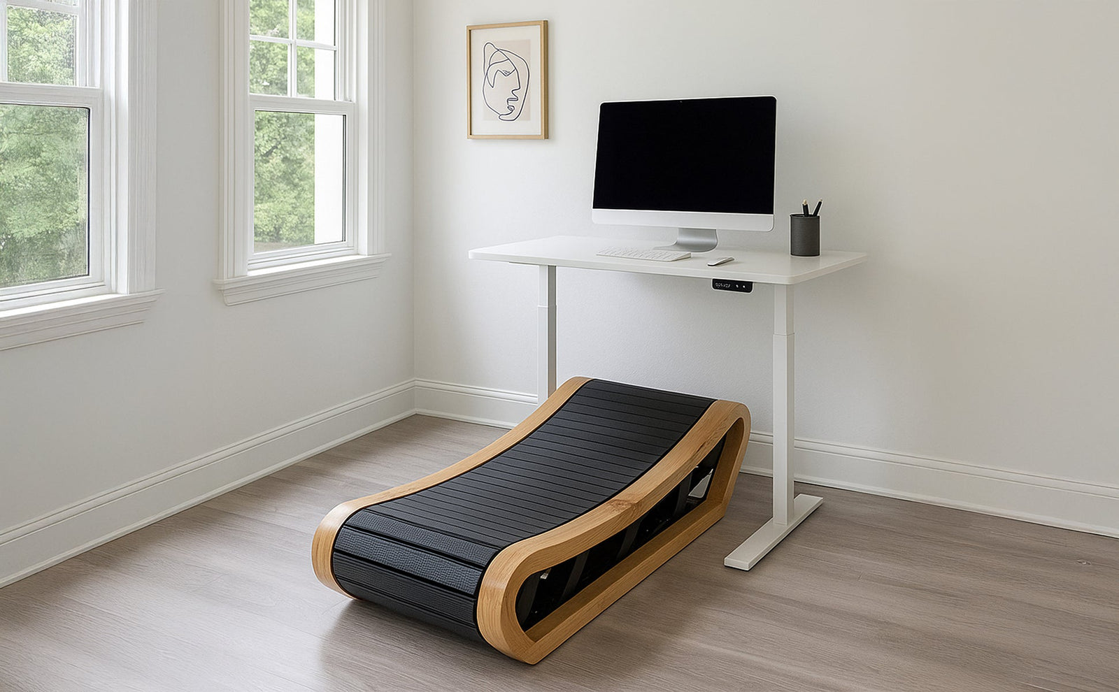 Ein gebogenes Holzlaufband mit schwarzem Laufbelag steht neben einem modernen Schreibtisch und einem Computer in einem hellen Raum mit großen Fenstern
