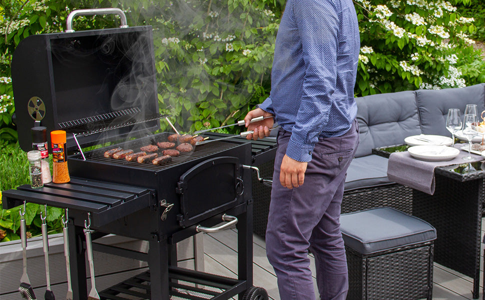 Ein tragbarer Holzkohlegrill mit rauchenden Steaks, umgeben von grünen Pflanzen und einer modernen Terrasse mit Sitzgelegenheiten und Tischgedeck