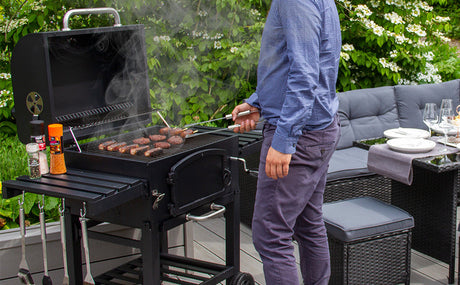 Ein tragbarer Holzkohlegrill mit rauchenden Steaks, umgeben von grünen Pflanzen und einer modernen Terrasse mit Sitzgelegenheiten und Tischgedeck
