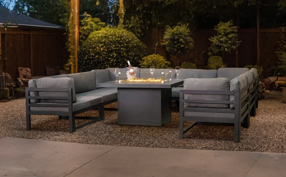 Moderne Gartenlounge mit grauem Polster, quadratischem Tisch und stimmungsvollem Licht, umgeben von grünen Pflanzen und Kiesboden