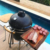 Schwarzer Kamado Holzkohlegrill mit glänzender Oberfläche, neben einem Holzbrett mit frischem Fleisch, vor einem blauen Pool und grünem Hintergrund