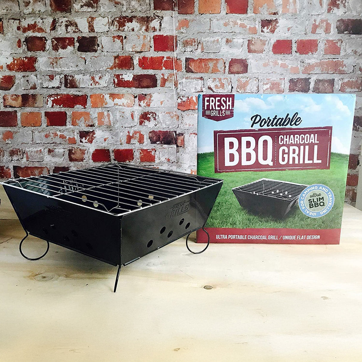 Tragbarer Grill in Schwarz mit verchromtem Grillrost, umgeben von einer rustikalen Ziegelwand und einer bunten Verpackung mit BBQ-Logo