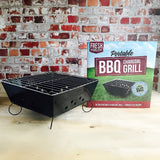 Tragbarer Grill mit schwarzem Metallgehäuse, verchromtem Grillrost und Kirschholz-Holzchips, präsentiert vor einer Backsteinwand und Verpackung