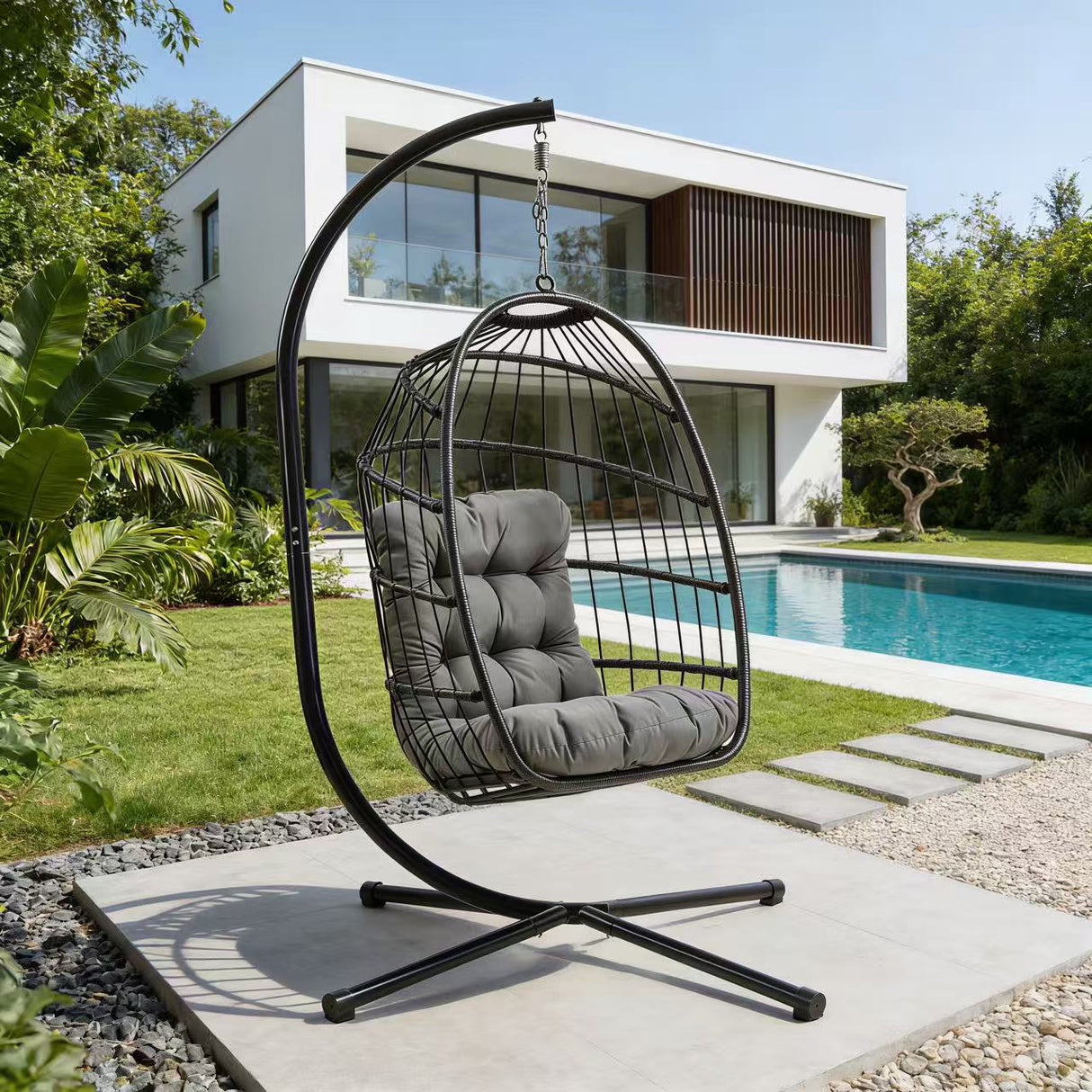 Schwarzer Rattan Hängesessel in Eiform mit grauem Kissen, elegant aufgehängt an einem stabilen Gestell, umgeben von einem modernen Garten und Pool