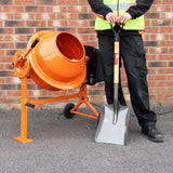 Orange Betonmischer mit runder Trommel, auf einem stabilen Gestell, neben einem Arbeiter in reflektierender Weste mit Schaufel in der Hand
