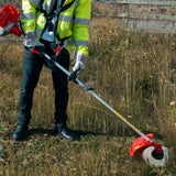 Multifunktionswerkzeug in leuchtendem Gelb und Rot, mit einem langen, stabilen Griff, der von einer Person in Arbeitskleidung gehalten wird, umgeben von hohem Gras