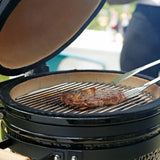 Schwarzer Kamado Holzkohlegrill mit geöffnetem Deckel, auf dem ein saftiges Steak auf den Grillrosten brutzelt, umgeben von einem eleganten Ambiente