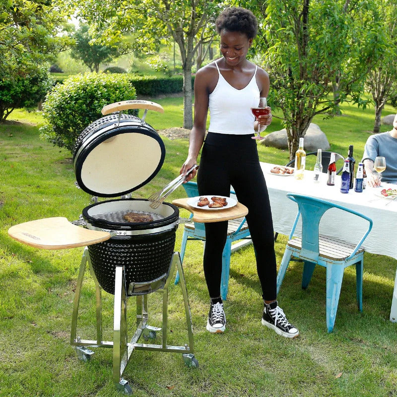 Schwarzer Kamado Holzkohlegrill mit Holzablagen, im Freien genutzt, während eine Person mit Grillzange Fleisch zubereitet, umgeben von Grün und Tischdeko