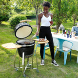 Schwarzer Kamado Holzkohlegrill mit Holzablagen, im Freien genutzt, während eine Person mit Grillzange Fleisch zubereitet, umgeben von Grün und Tischdeko