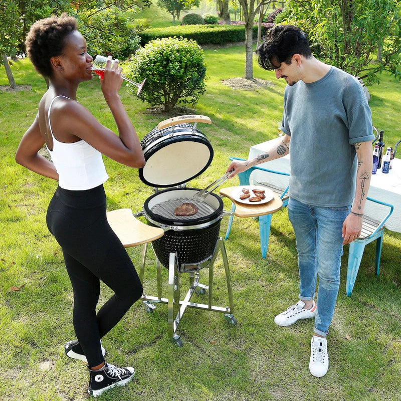 Ein 21-Zoll Kamado Holzkohlegrill in Schwarz, umgeben von frischem Gras, während zwei Personen beim Grillen von Steaks und Genießen eines Getränks sind