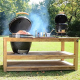 Kamado Holzkohlegrill in Schwarz auf einem Holzuntergestell, mit rauchendem Grilldeckel und zubereitetem Fleisch, umgeben von grünem Gras