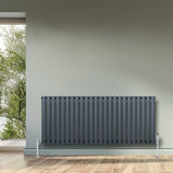 Gliederheizkörper in Anthrazitgrau, 600mm x 1440mm, mit vertikalen Lamellen und glänzenden Füßen, vor einer hellen Wand und Fenster mit Blick ins Grüne