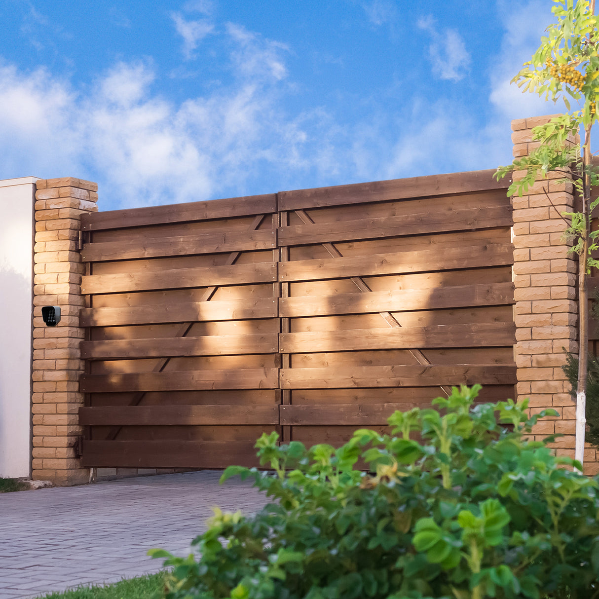 Holzschwingtor mit drahtloser Tastatur, rustikales Design, braune Holzlatten, umgeben von grünem Strauch und blauer Himmel im Hintergrund